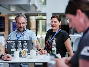 Two conference attendees wearing ID badges sit at a table during a professional event, smiling while engaging with others.