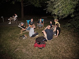 Group of people sitting in chairs and on ground in circle at night outdoor gathering in wooded area