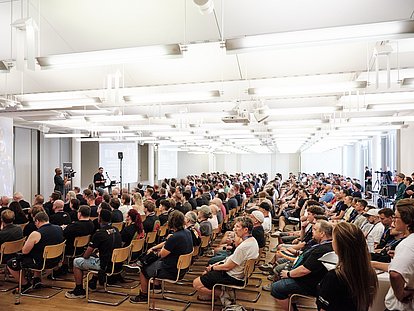 Large conference hall with hundreds of seated attendees listening to speakers on stage under bright fluorescent lighting