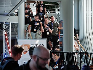 Group of people wearing lanyards and black shirts at a tech conference or event, standing on an elevated platform