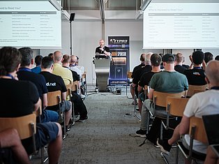 Speaker presenting at tech conference podium to seated audience with projection screens displaying content