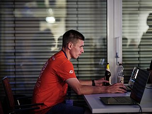 Man in red shirt working at laptop at desk with beverages and modern office background