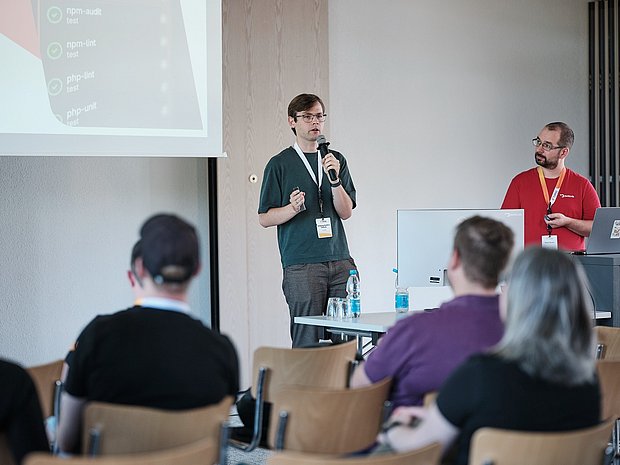 Speaker holding a microphone presenting to an audience at TYPO3 Developer Days, with another presenter standing by a laptop.