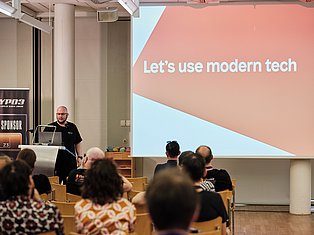 Speaker presenting at conference with slide reading 'Let's use modern tech' to seated audience in lecture hall