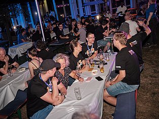 Large outdoor evening event with people seated at tables with drinks and food, blue and purple lighting, casual social gathering