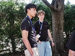 Two young men wearing lanyards stand outdoors among trees, both in black shirts, looking to the side.