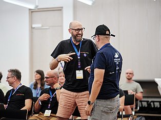 Man with glasses and lanyard speaking animatedly to another man at a conference or event in a bright room with other attendees.