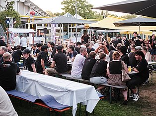 Large outdoor event with many people seated at tables under umbrellas and shade structures in a festive setting.