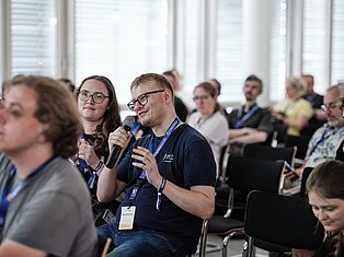 Man with microphone speaking at conference with audience members wearing ID badges in bright office setting