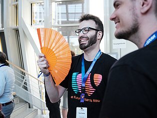 Man with glasses holding orange fan at conference, wearing lanyard badge, smiling while talking to colleague