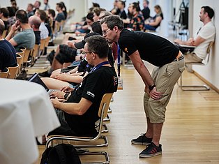 Instructor speaking with participants at a large conference or training event in a gymnasium with rows of seated attendees.