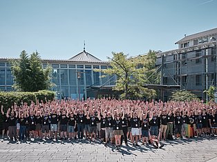 Large group of people with raised hands posing for photo in modern courtyard with contemporary buildings