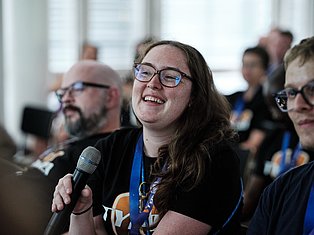 Woman with glasses speaking into microphone at conference, wearing blue lanyard badge, smiling at audience