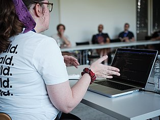 Person wearing glasses and purple headscarf presenting code on laptop to audience in conference room
