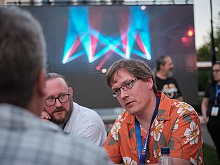 Man in orange shirt with glasses speaking at outdoor event with neon stage display behind