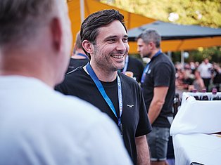 Smiling man wearing blue lanyard at outdoor event with crowd and orange building in background