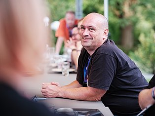Man in black polo shirt with lanyard smiling at outdoor garden gathering with people in background