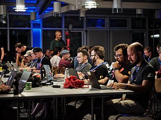 Group of developers and tech professionals working at laptops during a hackathon or tech conference event in a modern venue with blue lighting.