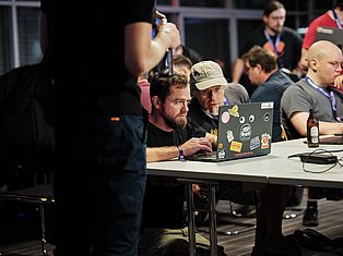 Man with beard working on sticker-covered laptop at tech conference panel with attendees and beer bottle