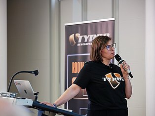 Woman wearing glasses and Tyro branded shirt speaking into microphone at podium with laptop and banner backdrop
