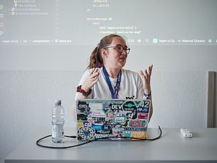 Developer presenting code on stage with sticker-covered laptop and conference badge