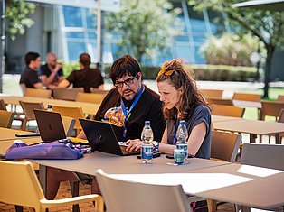 Two conference attendees with lanyards work together on laptops at an outdoor table in a modern campus setting.