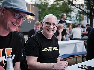Two men smiling at an outdoor community event, one wearing a cap and the other wearing glasses and a Sleaford Mods shirt