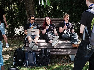 Three people wearing lanyards sit on a bench in a forest, one holding a laptop, surrounded by backpacks and other attendees.