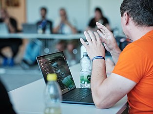 Man in orange shirt presenting at laptop during business meeting with blurred audience in background