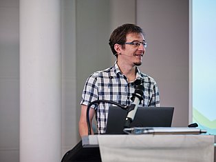 Man wearing glasses and checkered shirt speaking at podium with microphone and laptop