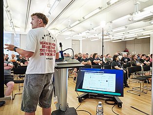 Man presenting at conference with laptop and display screen showing representation engineering diagram to seated audience in bright auditorium.
