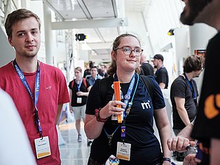 Conference attendees wearing ID badges stand in a bright modern venue, with a woman in glasses holding an orange object in the center.