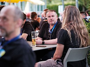 Man wearing blue lanyard at outdoor event table conversing with woman, holding drink glass surrounded by attendees