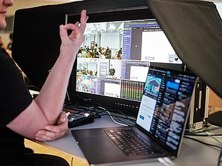 Person gesturing at video editing setup with multiple monitors displaying conference footage and editing software