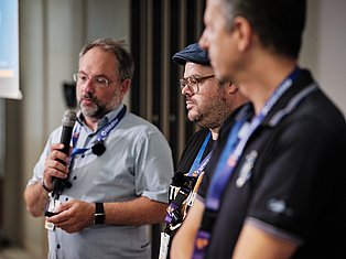 Three men with conference badges speaking at an event, one holding a microphone