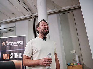 Man speaking at conference with microphone, holding water bottle, smiling in modern venue