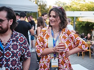 Woman in orange patterned shirt wearing event badge smiles at outdoor conference gathering with attendees