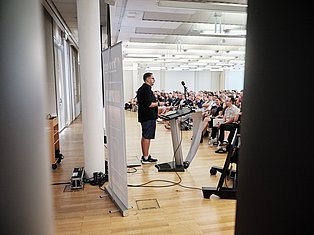 Man speaking at podium with microphone to large seated audience in bright modern conference hall with wooden floor