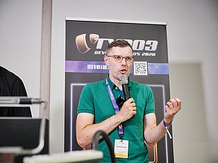Man in green polo shirt speaking at microphone during tech conference presentation with branded backdrop