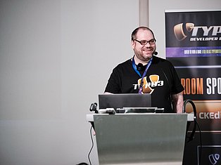 Man speaking at podium with laptop wearing headset and Typo3 shirt at developer conference
