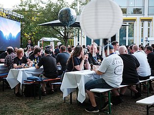 Outdoor event with large crowd seated at tables with white tablecloths, decorative balloons, and modern building in background