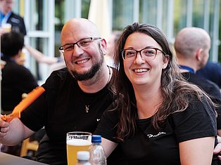 Two smiling people wearing glasses at an outdoor event, holding drinks and enjoying themselves with others in the background.
