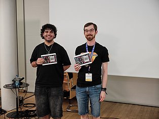 Two men holding 2021 event badges stand in front of a whiteboard in an indoor venue