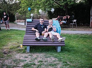 Two people sit on an oversized bench in a park, holding cameras, with decorative balloons and other visitors in the background.