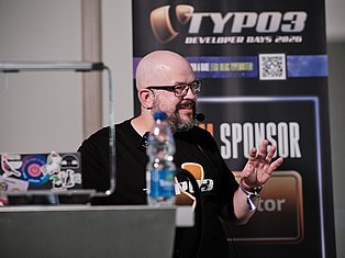 Bald man with glasses speaking at TYPO3 Developer Days 2026 conference with microphone and water bottle