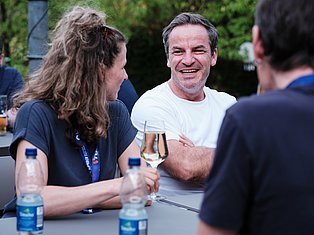 Man in white shirt laughing while conversing with woman and others at outdoor social gathering with drinks