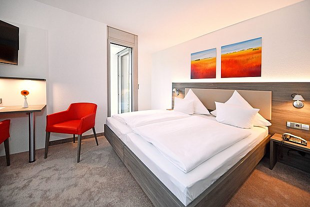 Bright hotel room with a double bed, white bedding, bedside tables with lamps, a small desk, and a red armchair near a window.