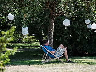 Man relaxing in deck chair under trees with hanging white paper lanterns in garden