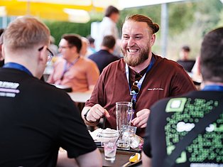 Man with beard wearing maroon shirt and lanyard smiling while conversing with others at outdoor event