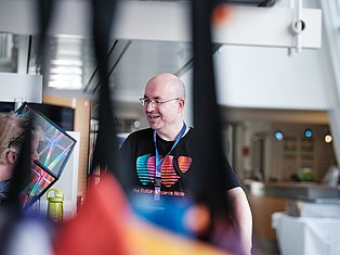 Man wearing glasses and conference badge speaking at tech event with colorful display screens